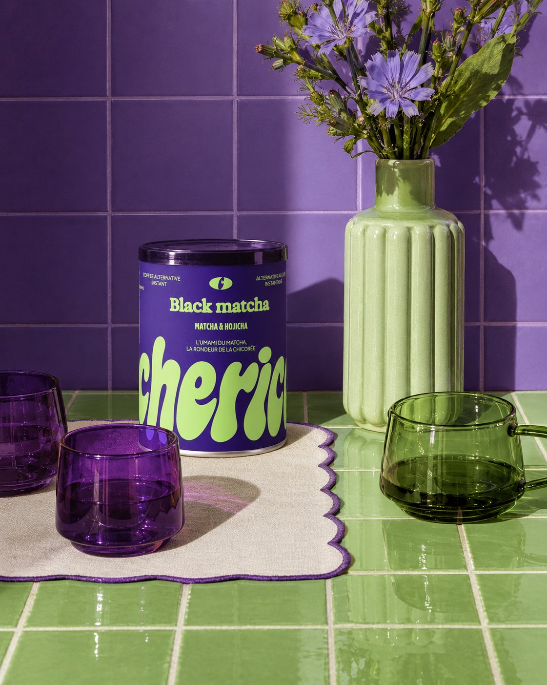 Pot Black Matcha Cherico sur un comptoir en carrelage vert, devant un mur violet, avec un vase de fleurs de chicorée